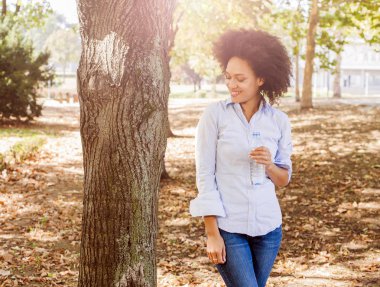 Gülümseyen genç siyah kadın portresi şişe su Doğa, afro saç stili, aşınma mavi gömlek, güneş ışığı fişek arka planda tutar