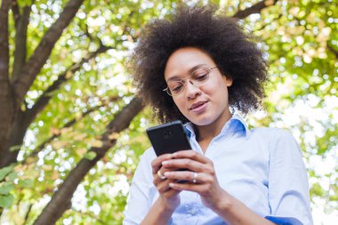 Telefonunuzun ve doğada güzel genç Afrikalı kadın. Açık portre seyir telefon gözlük, giyer