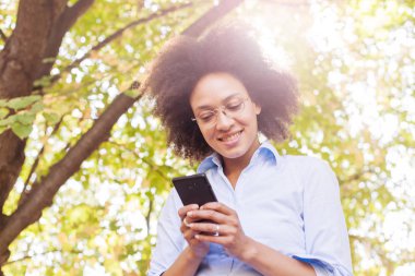 Güzel genç doğada telefon kullanan kadın siyah. Açık portre, giyer gözlük, telefon, güneş ışığı fişek arka planda bakarak