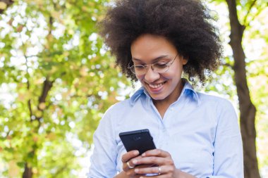 Telefonunuzun ve doğada güzel genç Afrikalı kadın. Açık portre seyir telefon gözlük, giyer