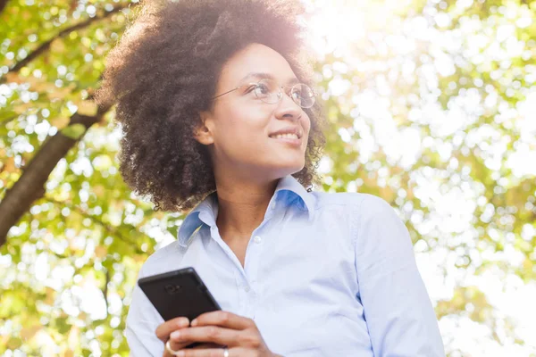 在大自然中使用智能手机的开朗年轻的黑人妇女 户外人像 戴眼镜 看着侧面 阳光在背景下闪光 — 图库照片