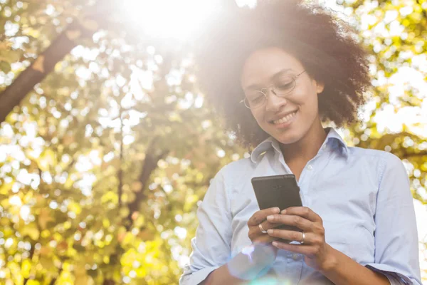 美丽的年轻黑人妇女使用手机在自然界中 户外人像 戴眼镜 看着电话 阳光在背景下闪光 — 图库照片