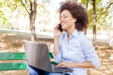 Dizüstü bilgisayarla çalışan ve telefon kullanan güzel melez bir kadın. Parkta bankta oturuyor, açık havada portre çiziyor, günlük kıyafetler ve gözlük takıyor. 