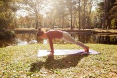 Young woman practising yoga on exercise mat in park near lake at beautiful morning autumn day.
