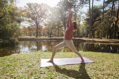 Doğada sağlıklı bir yaşam tarzı, güzel bir sonbahar sabahında gölün kenarındaki parkta yoga egzersizi yapan bir kadın..