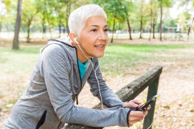 Sağlıklı kadın kıdemli ormanda koşu sonra telefon ile müzik dinleme sakin ol. Yaşam tarzı yetişkin insanlar egzersiz açık uygun.