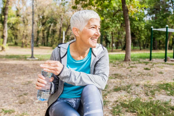 健康的老年妇女在户外健身公园锻炼后放松和保持一瓶水 — 图库照片