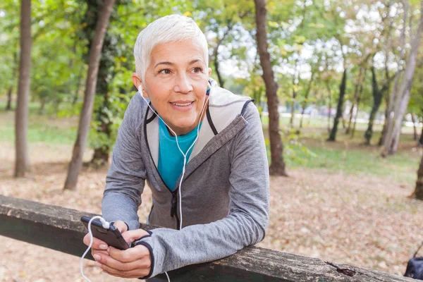 健康な年配の女性は フォレストでジョギングした後の携帯電話で音楽を聴くをリラックスします フィット ライフ スタイルの成熟した人が屋外トレーニング — ストック写真