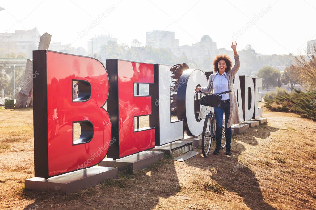 Happy traveler mixed race woman with rent bike wishes you a welcome in Belgrade, Sebia, Europe.