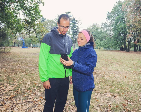 Fitness young woman show to sporty man on the smartphone, plan for exercising in forest. Sporty woman and men prepare for workout.