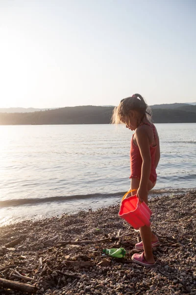 Vista posteriore della bambina che gioca sulla riva del fiume al tramonto, riflesso della luce solare sull'acqua — Foto Stock