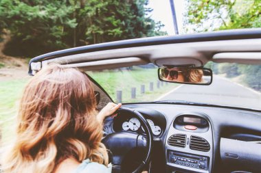 Yaz günü üstü açık araba kullanan güzel bir genç kadın. Güneş gözlüğü takıyordu. Arabanın dikiz aynasında yansıma var. Kadın sürücü arkası görünümü. Omuz üstü görünümü.