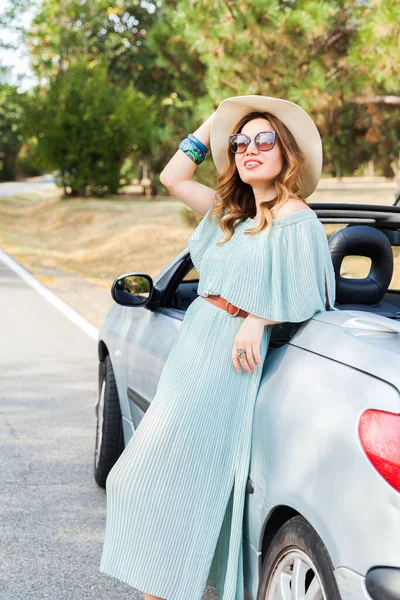 Attractive woman leaning and posing at convertible car. Happy pretty girl. Outdoor fashion portrait.Wearing elegant dress, straw hat and sunglasses.Looking at camera. Enjoying beautiful summer day in nature after driving car.