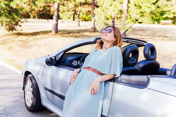 Outdoor fashion portrait. Attractive woman leaning and posing at convertible car. Happy pretty girl. Wearing elegant dress and sunglasses. Relaxing on summer day in nature