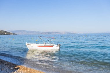 Küçük beyaz bir motorlu tekne, İyon Denizi 'nin turkuaz sularında, berrak mavi gökyüzünün altında sarı şamandıralar ve dağlık kıyı şeridi ile çevrili bir çakıl taşı plajı yakınında huzur içinde yüzüyor..