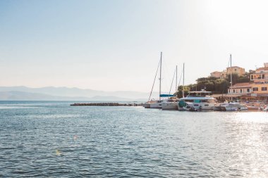 Huzurlu deniz manzarası, küçük bir Akdeniz yat limanında demirlemiş yelkenlileri ve lüks yatları, kıyı köyü ve parlak yaz güneşinin altında uzak puslu dağları yakalıyor..