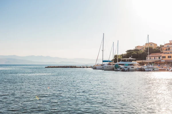 Huzurlu deniz manzarası, küçük bir Akdeniz yat limanında demirlemiş yelkenlileri ve lüks yatları, kıyı köyü ve parlak yaz güneşinin altında uzak puslu dağları yakalıyor..