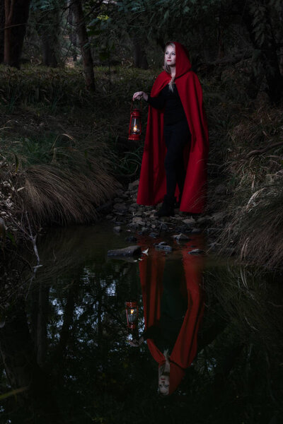 Attractive woman dressed a little red riding-hood cross a stream in a dark forest with a lantern