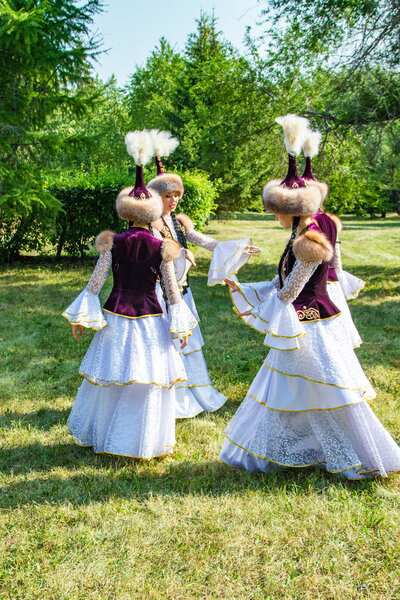 Petropavlovsk, Kazakhstan - August 17, 2019: Girls in national Kazakh clothes in hats. Summer day, green grass of a tree.