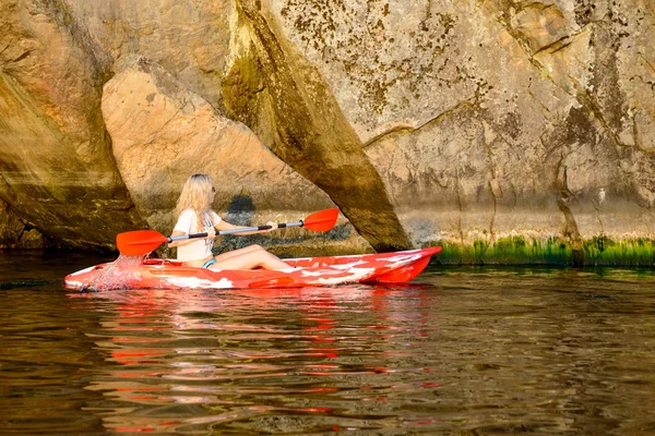 晚上在高岩石下的美丽的河或湖的年轻妇女划红色独木舟 — 图库照片