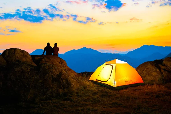 Young Couple Sitting on the Rock near Illuminated Tent and Watching the Beautiful Evening Mountain View with Sunset Sky — Stock Photo, Image