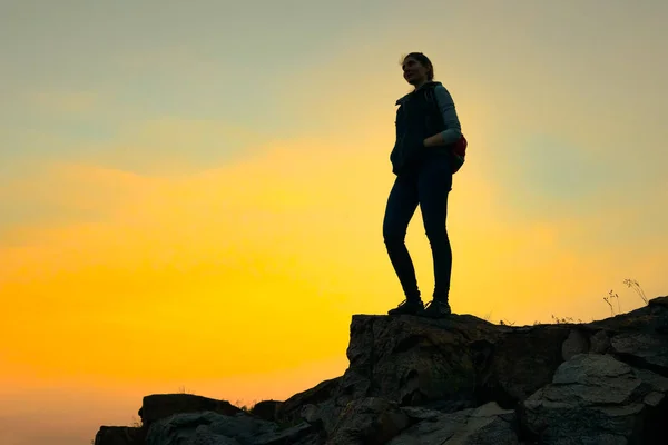 Νεαρή γυναίκα ταξιδιώτης με σακίδιο στέκεται στην κορυφή του βράχου στο καλοκαιρινό ηλιοβασίλεμα. Έννοια ταξιδιού και περιπέτειας. — Φωτογραφία Αρχείου