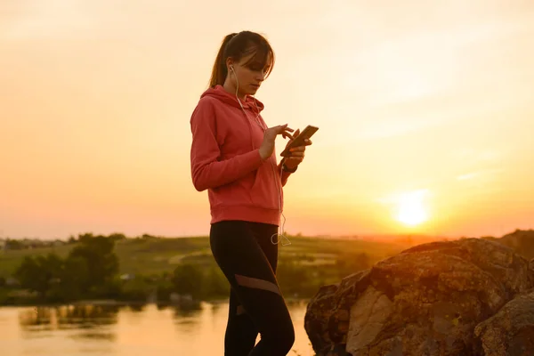 女跑步者在锻炼身体后休息，在落日落山时使用智能手机和听音乐。 体育概念. — 图库照片