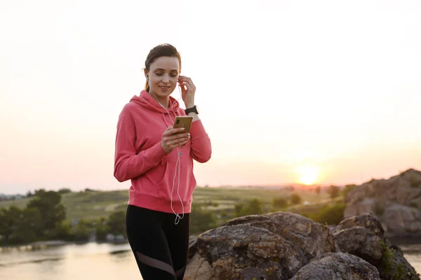 Žena Runner Odpočívá po cvičení, Použití Smartphone a poslech hudby při západu slunce na skále. Sportovní koncepce. — Stock fotografie