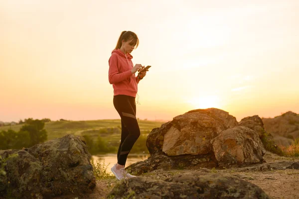 Woman Runner Descansando después del entrenamiento, usando Smartphone y escuchando música en Sunset on the Rock. Concepto deportivo . —  Fotos de Stock