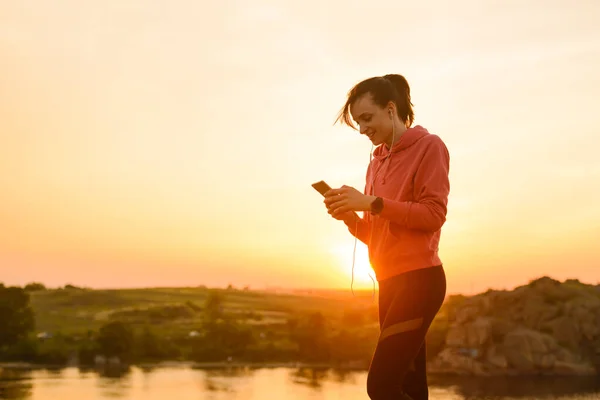 Woman Runner Descansando después del entrenamiento, usando Smartphone y escuchando música en Sunset on the Rock. Concepto deportivo . —  Fotos de Stock