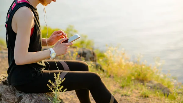 Ung kvinna löpare använder smartphone och lyssnar på musik vid solnedgången på berget spåret. Sportkoncept. — Stockfoto