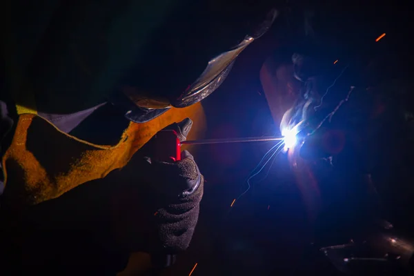 A welder at work. Sparks flying. Welder in mask at work. Profession of a welder. Sparks around. Welding metal. Dark background. Bright light. — Stock Photo, Image