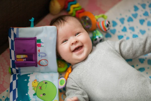 Ребенок в сером комбинезоне лежит среди развивающихся игрушек улыбаясь — стоковое фото