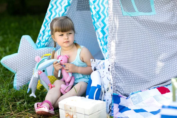 かわいい女の子がおもちゃを持って通りの夏にファブリックハウスに座っている ロイヤリティフリーのストック写真