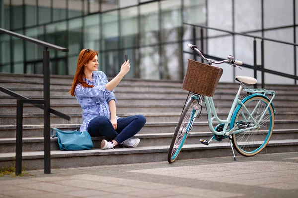 Woman sitting on stairs and making photo near bike in town