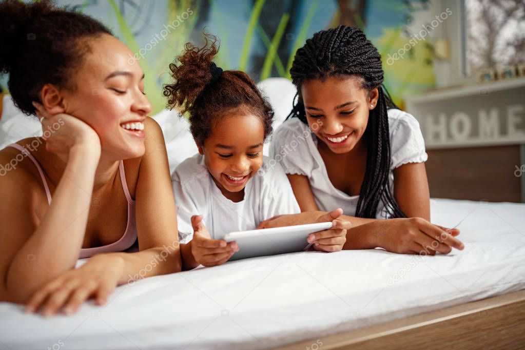 Family time-Smiling cute girl playing with mother and sister at home in a bed on digital tablet
