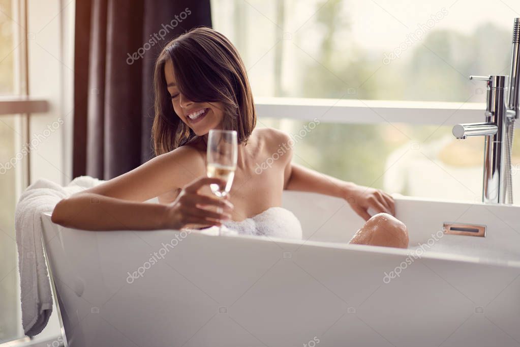 Smiling young woman relaxing in bath with foam and drink vine