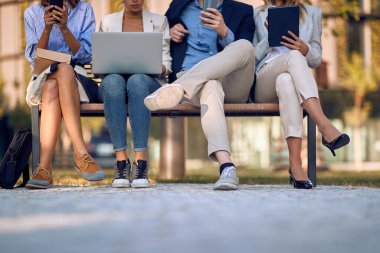 Açık hava bankında bacakları üzerinde oturan iş insanlarının cep telefonlarını, laptoplarını, tabletlerini kontrol eden kırpılmış görüntüleri. vücut dili kavramı