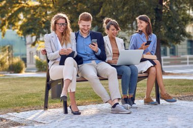 Genç insanlar parkta birlikte gülüyorlar, bankta oturuyorlar, cep telefonlarını, laptoplarını, tabletlerini tutuyorlar. İyi haberi ya da şakayı paylaşmak
