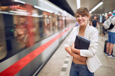 Metro istasyonunda elleri çaprazlanmış, gülümseyen, klasör ve defter tutan beyaz bir iş kadını. Vücut dili. Zaman kavramı