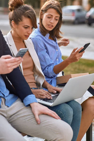 在公园里的笔记本电脑上打字的年轻成年女性坐在公园的长椅上 与朋友们一起使用手机 现代技术 — 图库照片