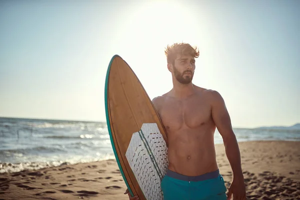 ハンサムでセクシーな若い白人男性が日当たりの良いビーチに立ってサーフボードを持って — ストック写真