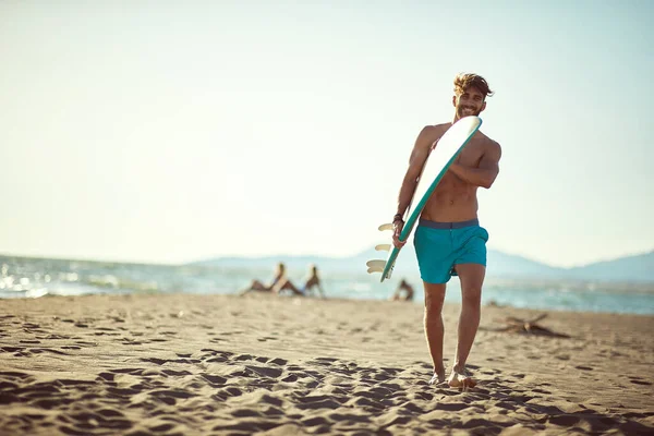 日当たりの良いビーチを歩きサーフボードを持って笑顔で若い白人のベアディ男性に合う — ストック写真