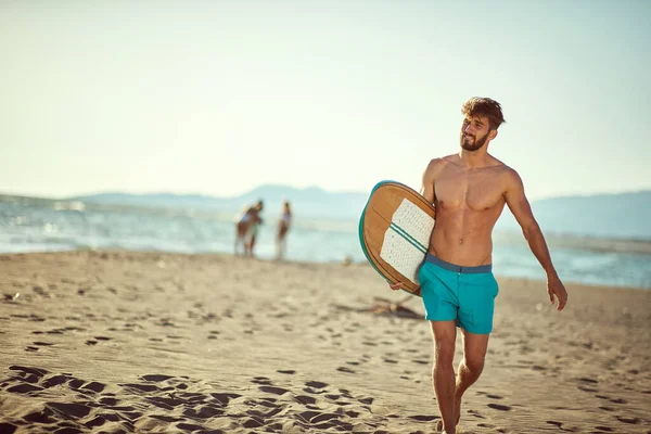 日当たりの良いビーチを歩く若い白人のベアディ男性に合うサーフボードを持ち笑顔で スペースのコピー — ストック写真
