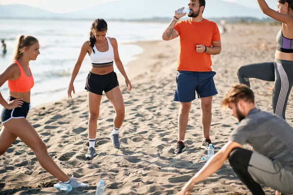 若い白人の大人たちが砂浜で運動をしています プラスチックボトルから水を飲むベアディ男 — ストック写真