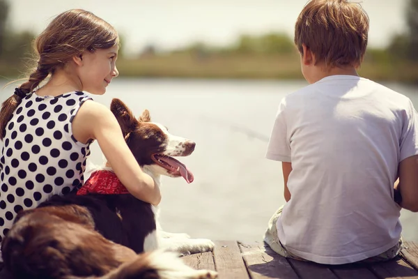 小さな男の子と女の子で犬が木の桟橋に座って池で釣りをしている — ストック写真