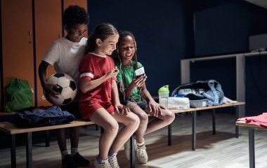 Gülümseyen çocuklar soyunma odasında oturuyorlar ve antrenmandan önce telefonla müzik dinliyorlar. .