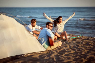 Bir grup arkadaş plajda şarkı söylüyor, günbatımının tadını çıkarıyor, gülümsüyor, gülüyor.