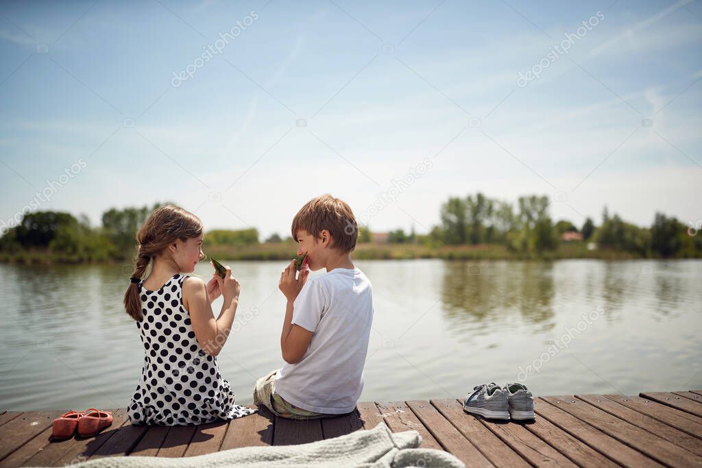 Smiling children  on wooden near pond eating fresh watermelon.