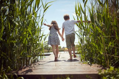 Erkek ve kız kardeş çıplak ayakla gölün rıhtımında yürüyorlar.